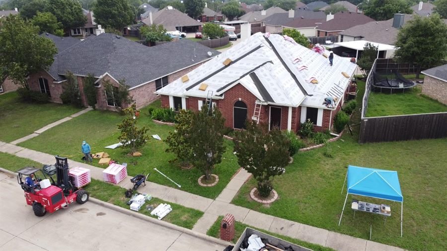 Drone view of a house fully covered in white synthetic underlayment with a crew working and shingle bundles staged at the kerb