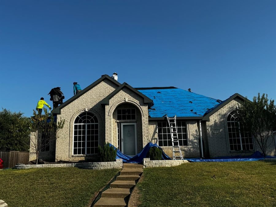 Front of a house with tarps laid over the landscaping and a crew removing shingles from the roof